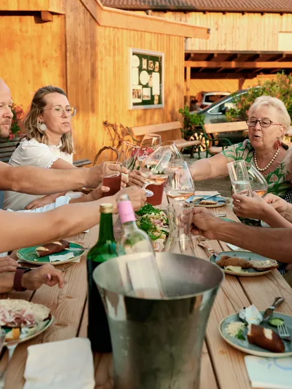 Family and friends toasting drinks at a wooden outdoor table with food in a rustic setting