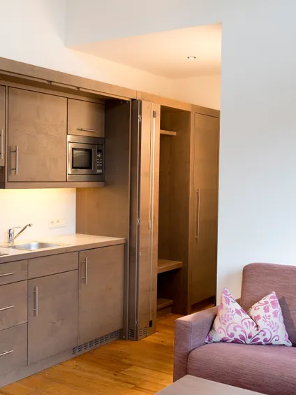 Kompakte Studioküchenzeile mit Holzschränken, Mikrowelle und lila Sofa mit Blumen-Kissen
