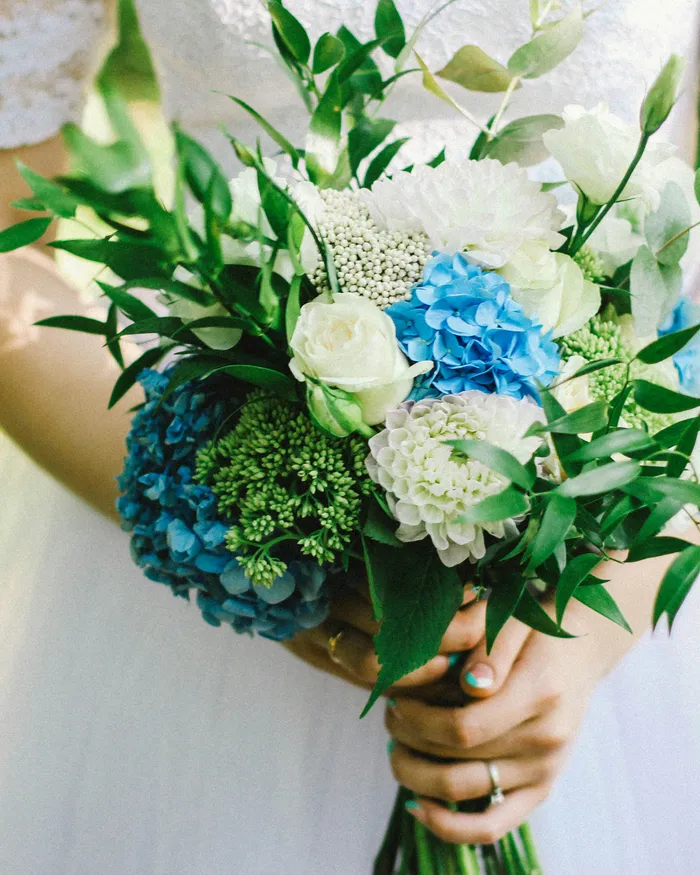 Braut im weißen Kleid mit einem Strauß aus blauen Hortensien, weißen Rosen und grünem Laub