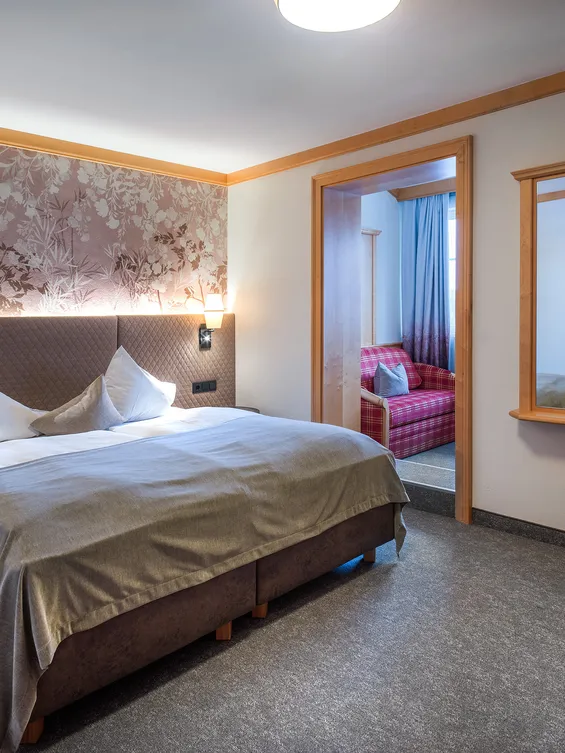 Gemütliches Hotelzimmer mit Doppelbett, floralem Kopfteil, Wandleuchten und angrenzender Sitzecke