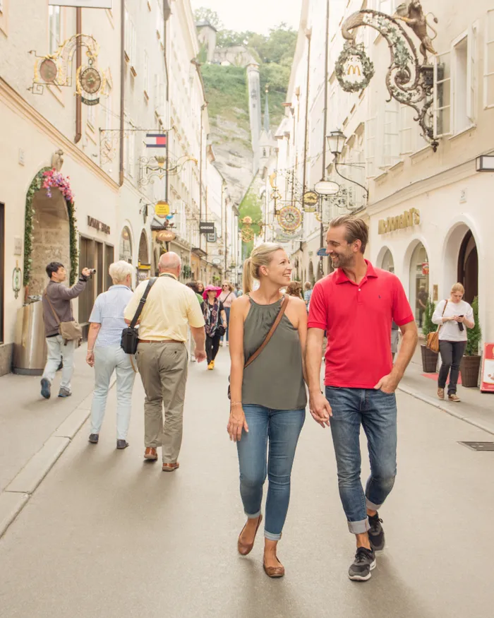 Paar spaziert durch belebte Einkaufsstraße in Salzburg mit historischen Schildern und Boutiquen
