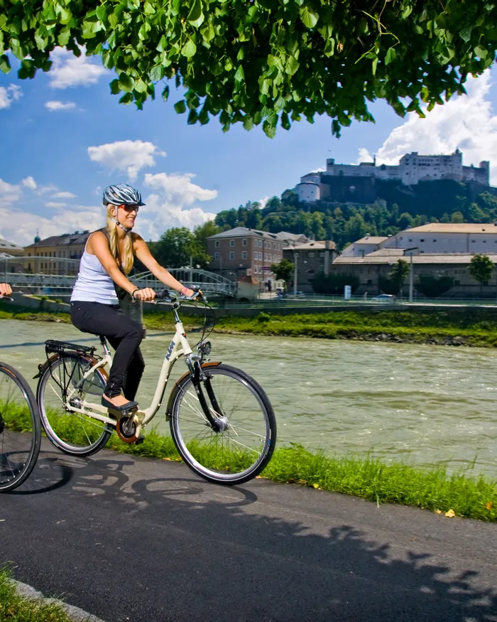 Coppia in bicicletta lungo il fiume Salzach con la fortezza Hohensalzburg sullo sfondo