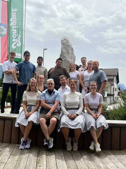 Hotelteam posiert gemeinsam in Tracht vor Steinmonument und Fahnen