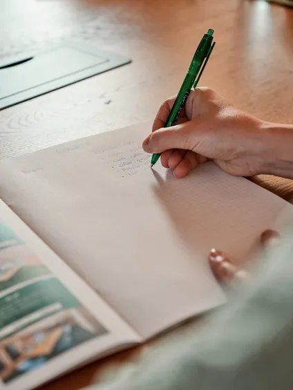 Person schreibt mit grünem Stift Notizen in ein Heft auf einem Holztisch