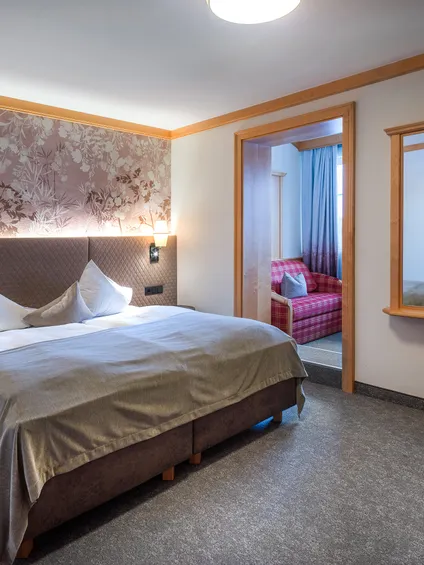 Gemütliches Hotelzimmer mit Doppelbett, floralem Kopfteil, Wandleuchten und angrenzender Sitzecke