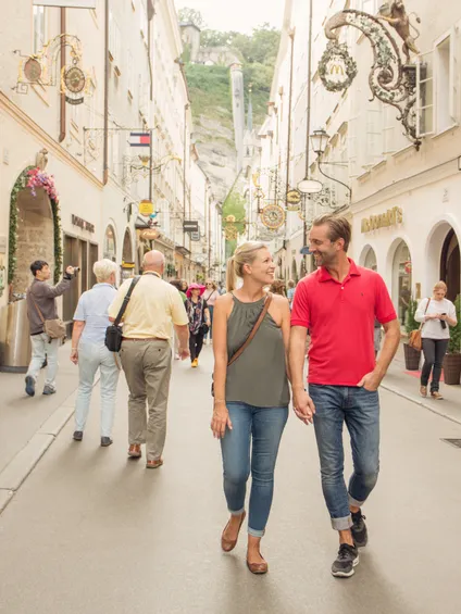 Paar spaziert durch belebte Einkaufsstraße in Salzburg mit historischen Schildern und Boutiquen