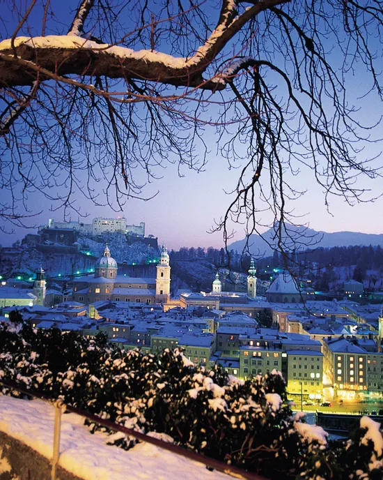 Snow-covered Salzburg cityscape at dusk with Hohensalzburg Fortress in the background