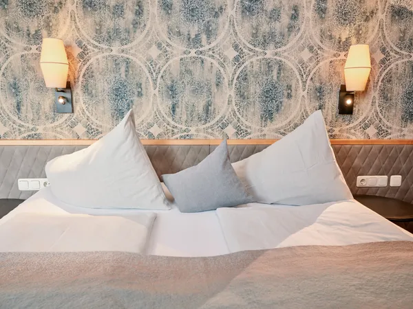 Gemütliches Hotelbett mit großen Kissen, gemusterter Tapete und warmen Wandleuchten
