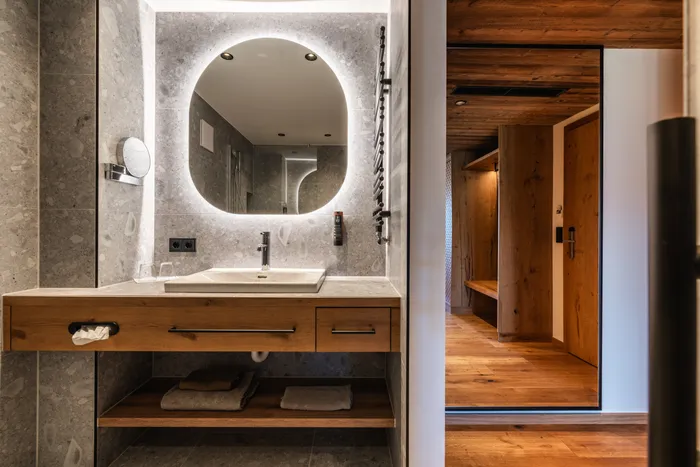 Modern bathroom with round backlit mirror, stone walls, and wooden vanity next to hallway