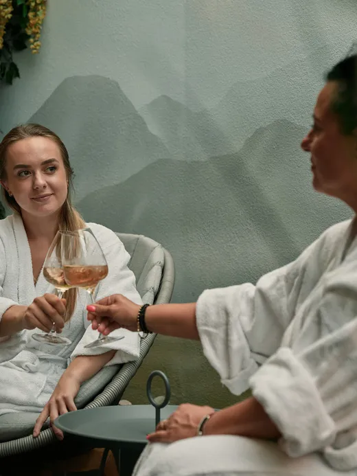 Zwei Frauen in Bademänteln stoßen mit Weingläsern in entspannter Wellness-Atmosphäre an