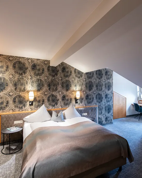 Modernes Hotelzimmer mit Doppelbett, gemusterter Tapete, Schreibtisch und gemütlicher Beleuchtung