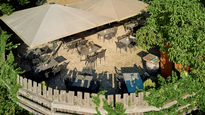 Outdoor restaurant terrace with empty tables under large beige umbrellas and surrounded by greenery