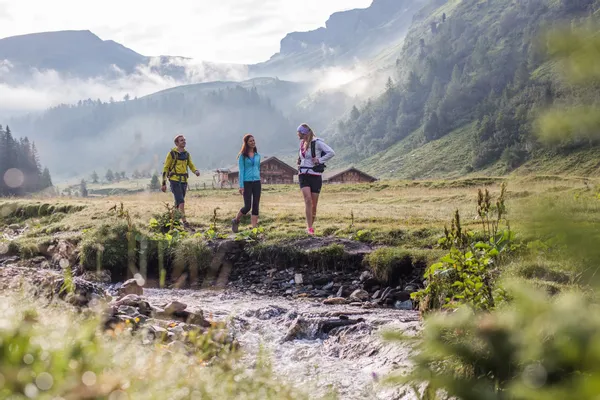 Wandergruppe läuft an einem Bach im Almtal mit Berghütten und nebelverhangenen Gipfeln entlang