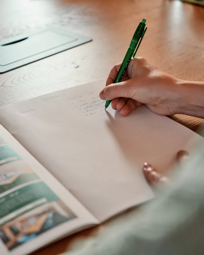 Person schreibt mit grünem Stift Notizen in ein Heft auf einem Holztisch