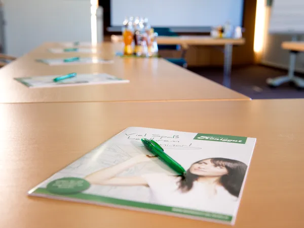 Sala seminari con tavoli in legno, brochure, penne verdi e bevande sullo sfondo