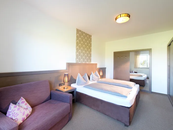 Helles Hotelzimmer mit Doppelbett, lila Sofa und großem Spiegelkleiderschrank