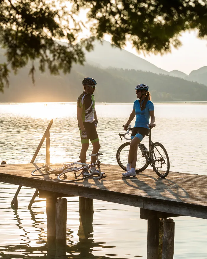 Ciclisti che parlano su un molo al lago al tramonto con paddleboarder sullo sfondo