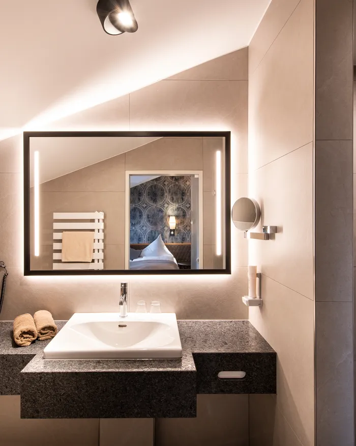 Modern hotel bathroom with illuminated mirror, sink, and view into a cozy bedroom