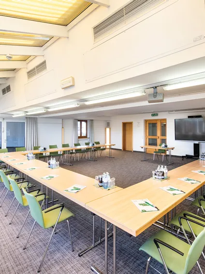 Heller Konferenzraum mit U-förmiger Tischanordnung, grünen Stühlen und großem Bildschirm vorne
