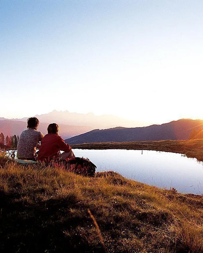 Paar sitzt bei Sonnenuntergang am Bergsee mit Alpen im Hintergrund