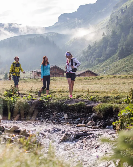 Wandergruppe läuft an einem Bach im Almtal mit Berghütten und nebelverhangenen Gipfeln entlang