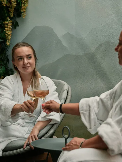 Zwei Frauen in Bademänteln stoßen mit Weingläsern in entspannter Wellness-Atmosphäre an