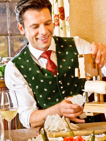 Mann in traditioneller bayerischer Tracht schneidet Käse an einem rustikalen Restauranttisch