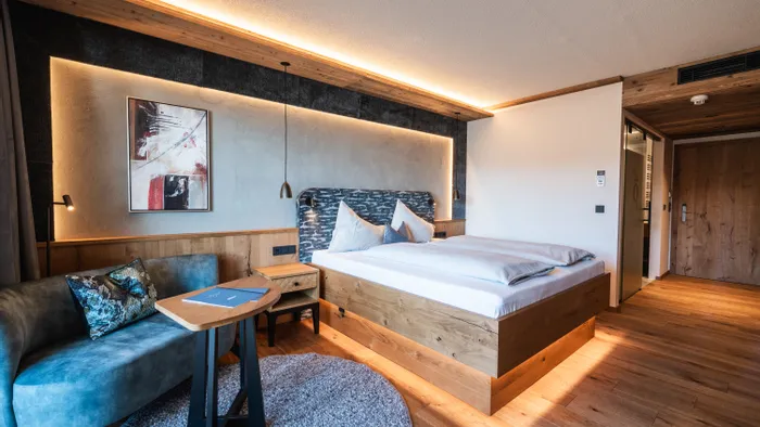 Modern hotel room with wooden flooring, large bed, blue sofa, and warm ambient lighting