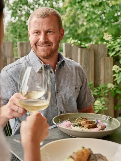 Mann lächelt und stößt mit Weißwein an einem Restauranttisch im Freien an