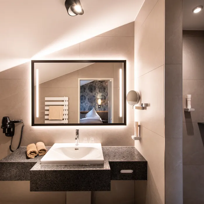Modernes Hotelbad mit beleuchtetem Spiegel, Waschbecken und Blick ins gemütliche Schlafzimmer