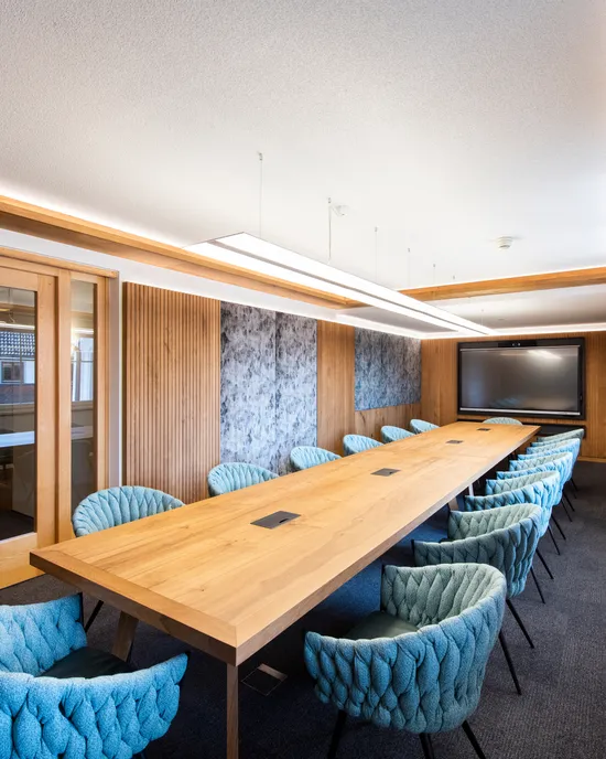 Moderner Konferenzraum mit langem Holztisch, blauen Stühlen und großem Bildschirm