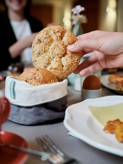 Hand greift nach Sesambrötchen aus Frühstückskorb am Tisch