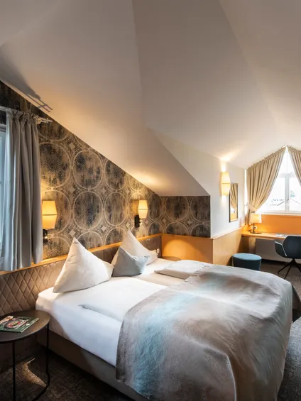 Gemütliches Dachgeschoss-Hotelzimmer mit Doppelbett, Schreibtisch, gemusterter Tapete und großen Fenstern