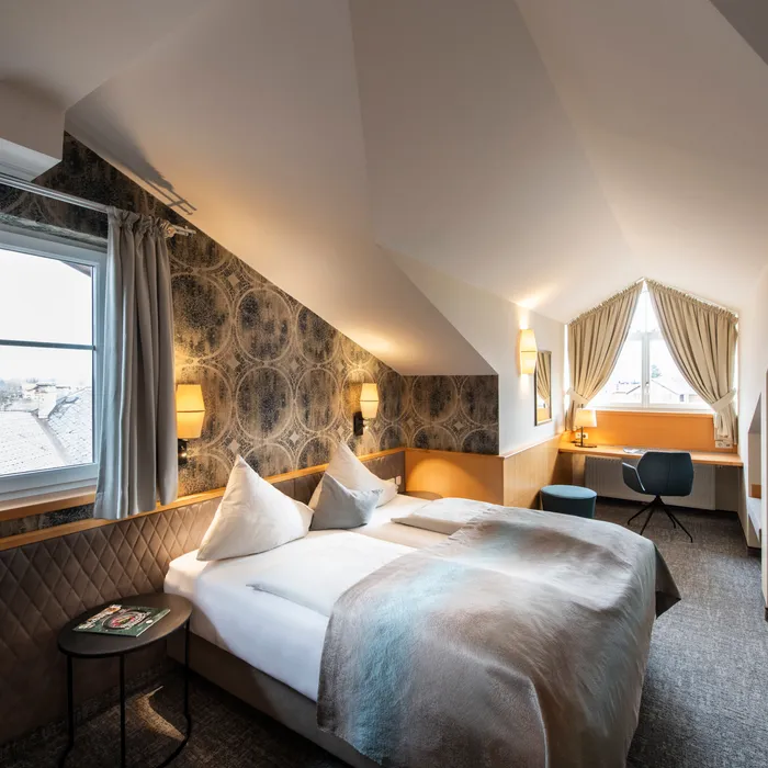 Gemütliches Dachgeschoss-Hotelzimmer mit Doppelbett, Schreibtisch, gemusterter Tapete und großen Fenstern