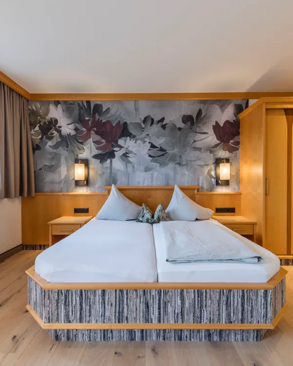 Hotelzimmer mit Holzmöbeln, Doppelbett, Blumen-Tapete und Fenster mit Vorhängen